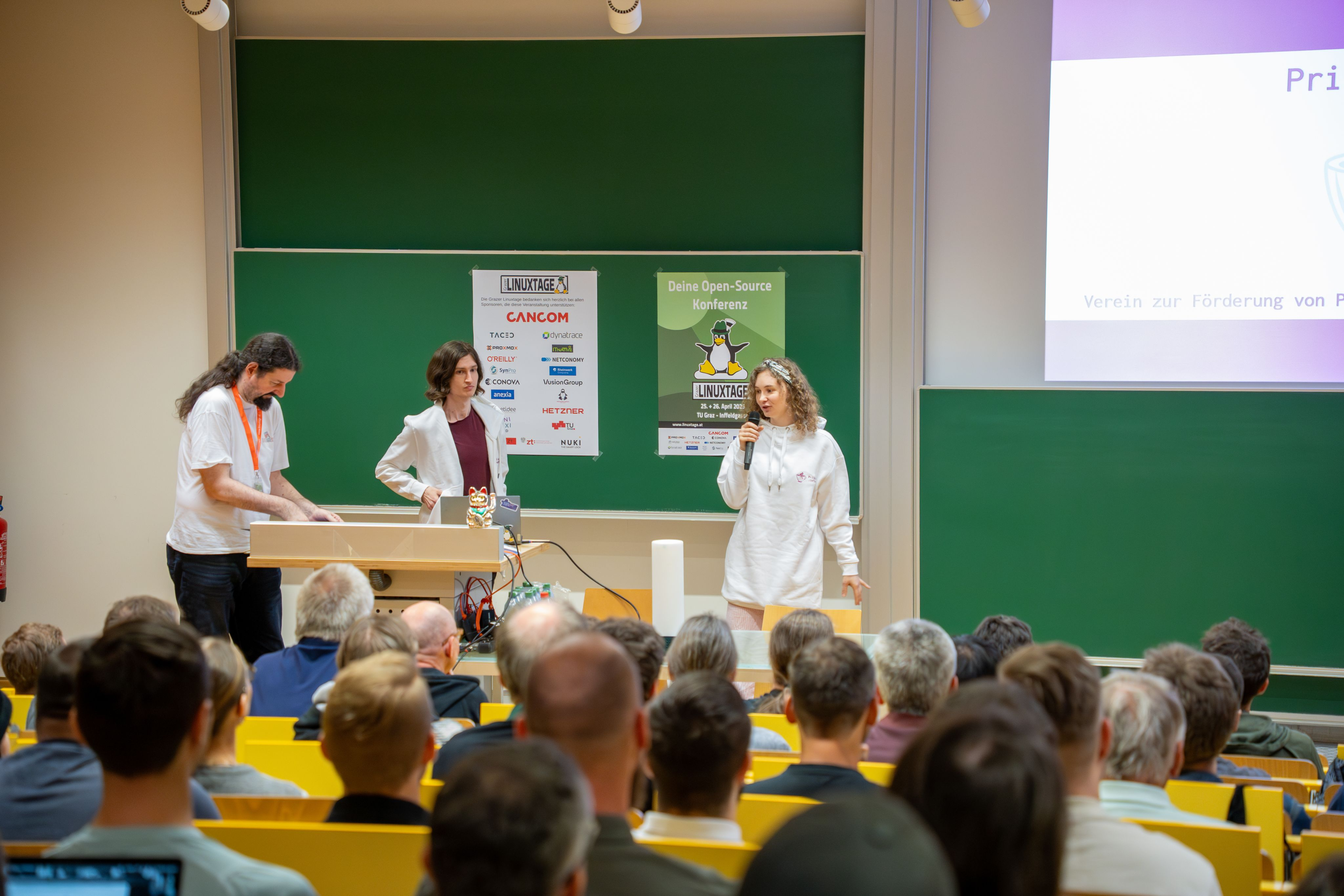 Jupfi at the lightning talk. A crowd is visible watching jupfi giving the lightning talk. The Linuxtage mascot (a penguin with a Steirerhut) is visible on a chalkboard.