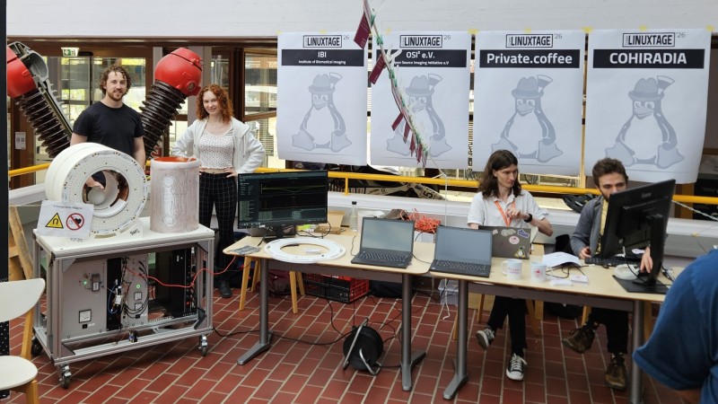 A view of the full booth. From left to right: a low-field MRI scanner with two people in the back, the institute of biomedical imaging booth, the open source imaging initiative booth, the private.coffee booth with two people being present and the COHIRADIA booth.